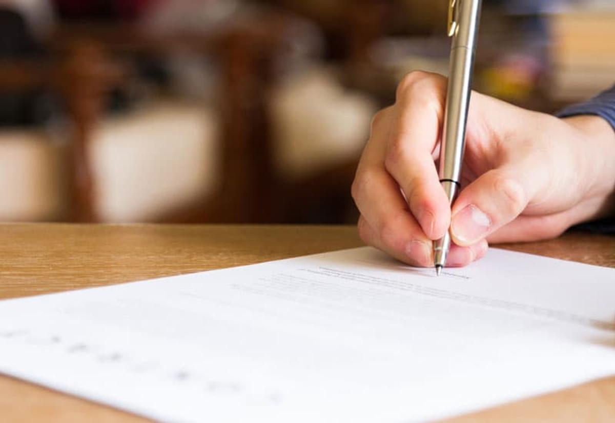 Close-up of a hand holding a pen and writing on a sheet of paper on a desk.