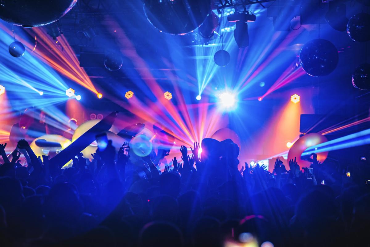 Crowd dancing at a lively nightclub concert with colorful stage lights, laser beams, and raised hands creating a high energy party atmosphere.