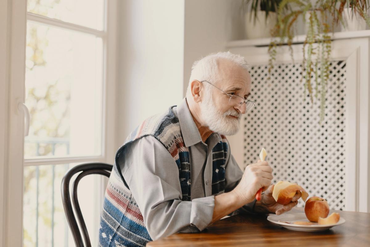 Older man sitting at a table peeling an apple, highlighting the role of balanced nutrition and healthy daily habits in recovery.