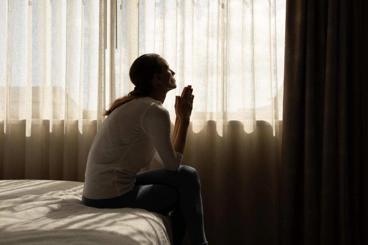 Woman sitting on a bed by a window with hands clasped, looking upward in quiet reflection.