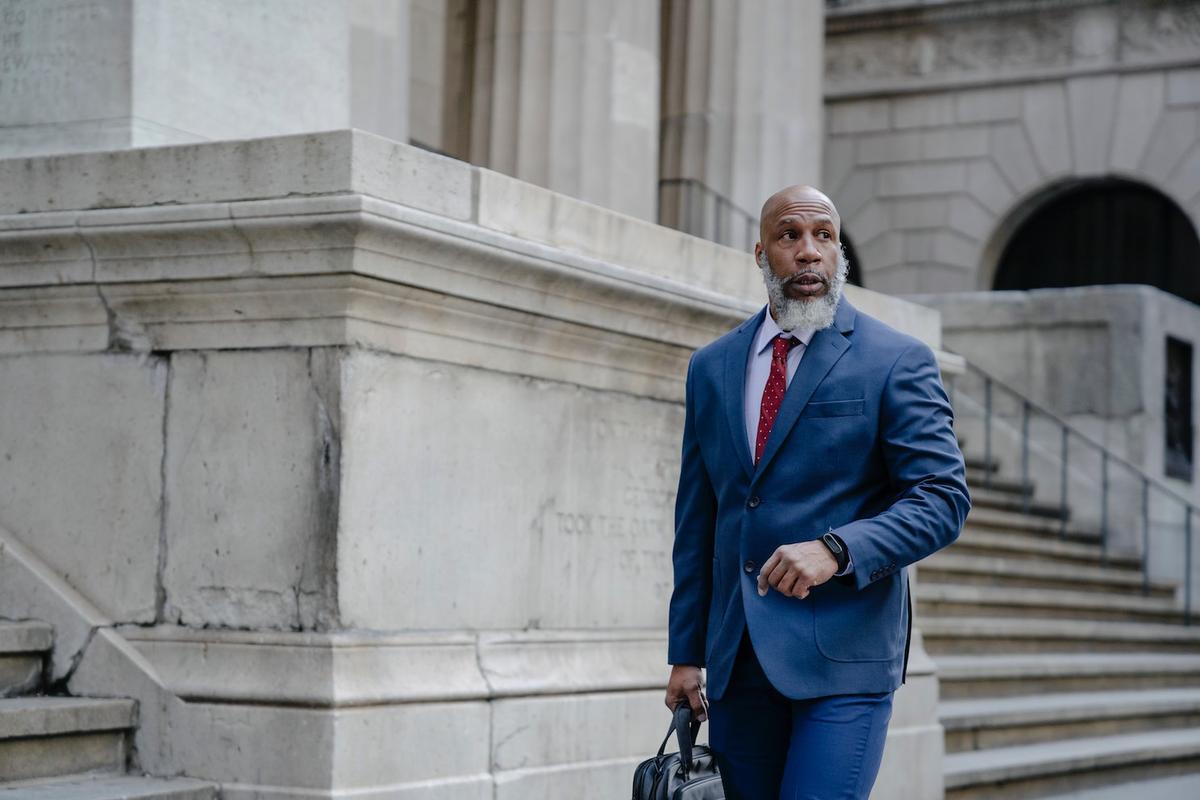 A well-dressed man in a blue suit walking confidently past a large stone building, representing social status in recovery and rebuilding professional life after addiction.