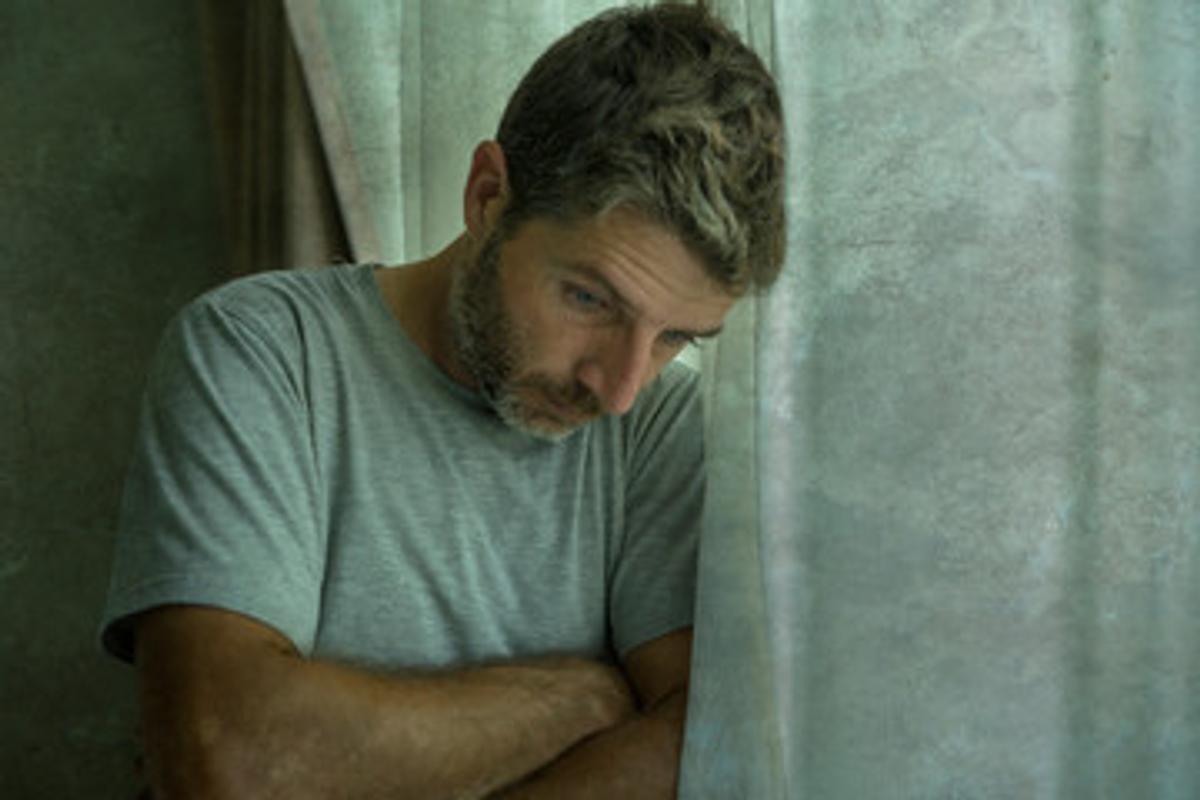 A middle-aged man with a beard and a gray t-shirt stands by a window, looking downcast with his arms crossed, conveying sadness, depression, or worry.