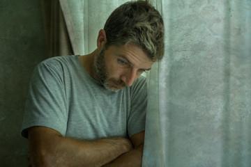 A middle-aged man with a beard and a gray t-shirt stands by a window, looking downcast with his arms crossed, conveying sadness, depression, or worry.