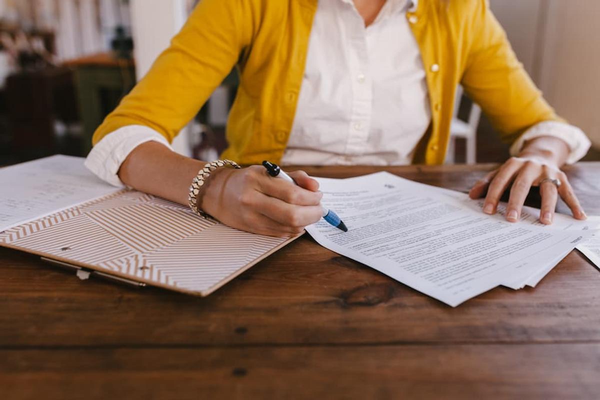 A person in a yellow cardigan and white shirt sitting at a wooden table, reviewing and pointing at a printed document with a pen.