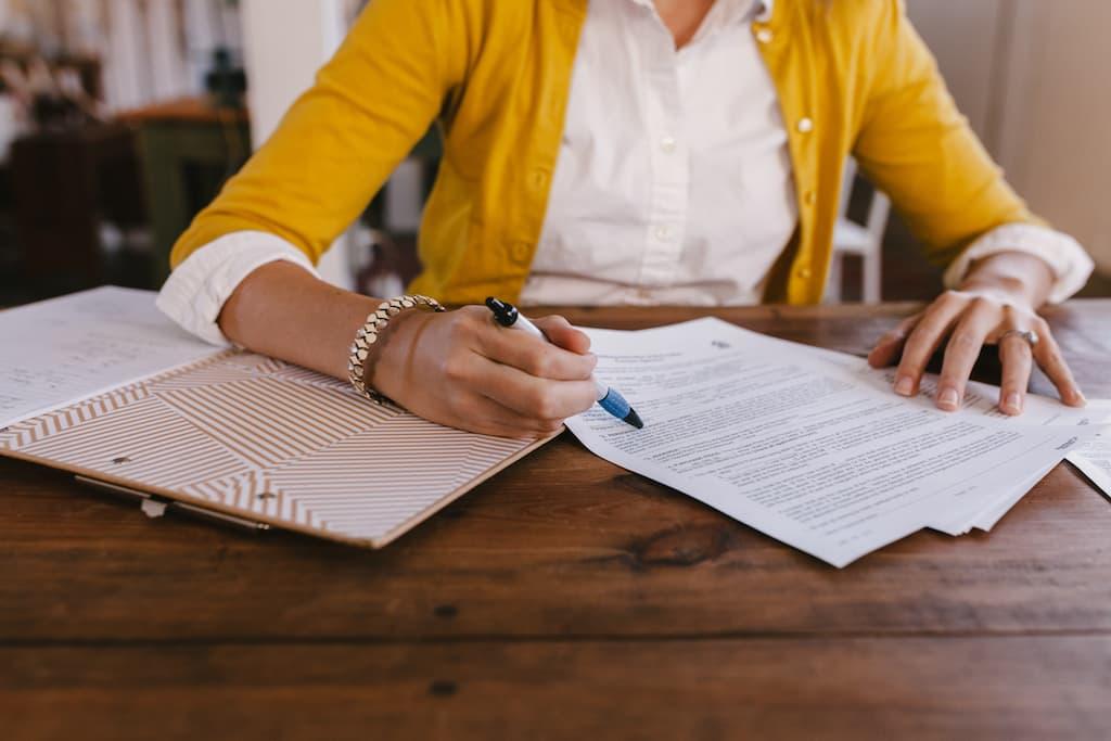 A person in a yellow cardigan and white shirt sitting at a wooden table, reviewing and pointing at a printed document with a pen.