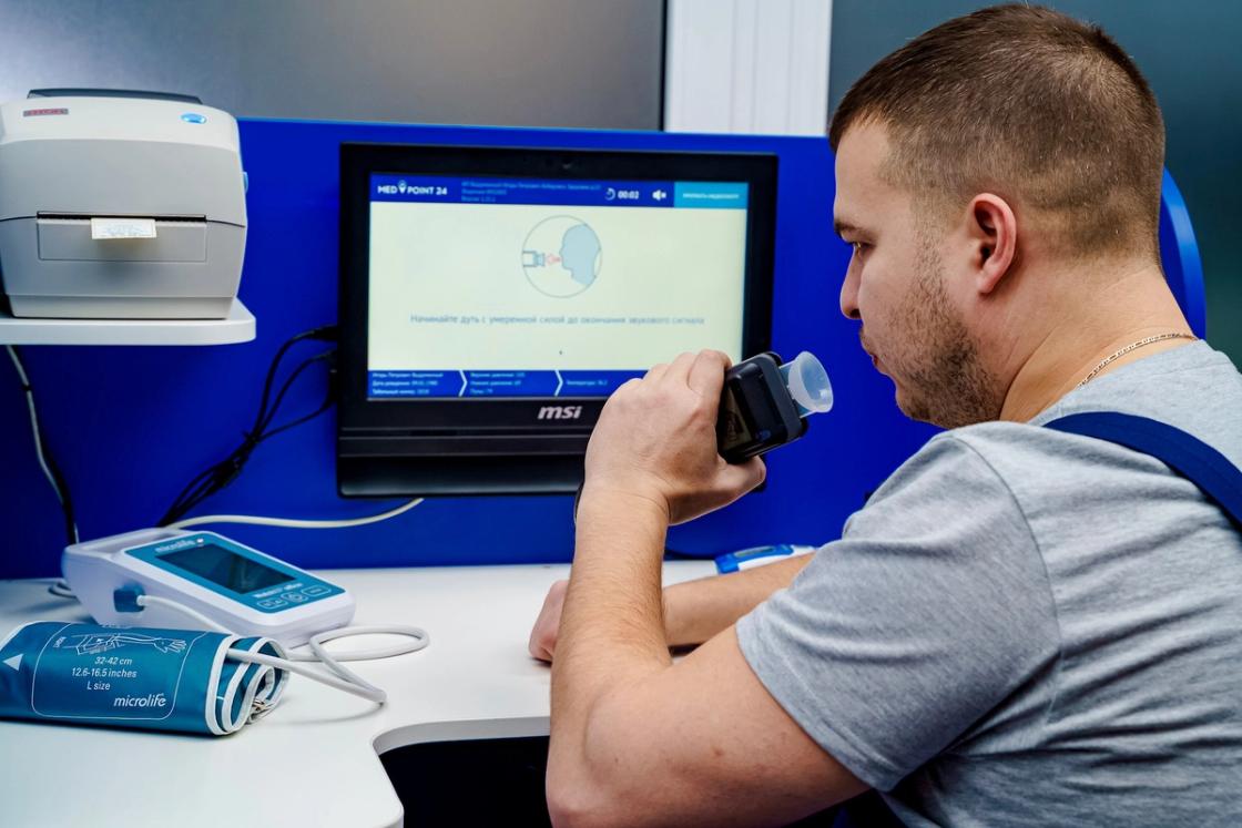 A man in a grey t-shirt sits at a medical station, breathing into a digital breathalyzer or spirometer.