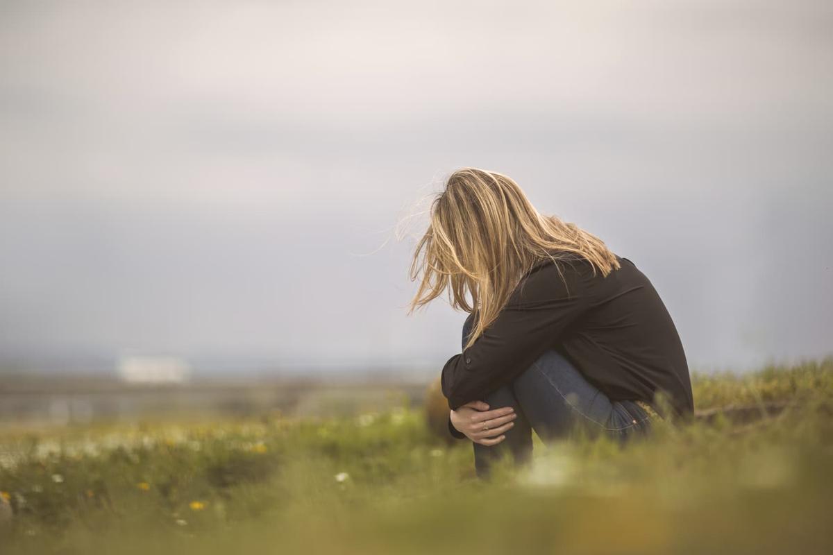 A woman sits alone on a grassy hill with her head bowed and her arms wrapped around her knees.