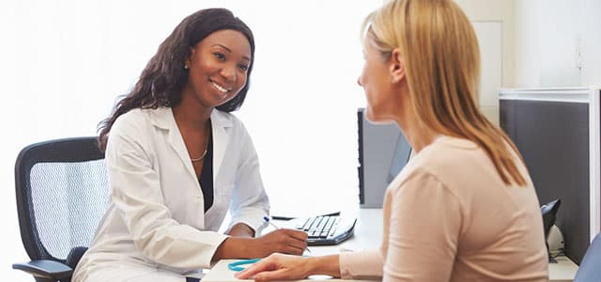 A smiling female doctor in a white lab coat sits at a desk, looking at and talking with a blonde patient during a consultation.