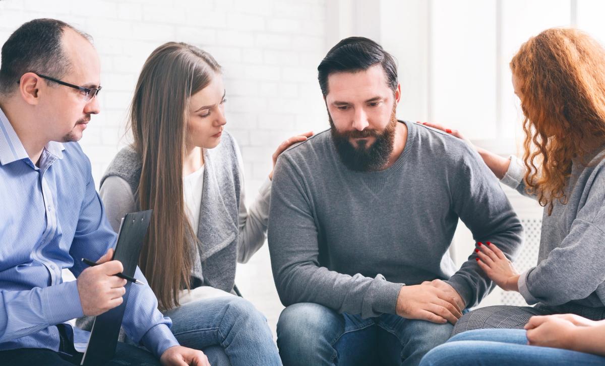 A group of people sitting in a circle during a support group session. Two women place comforting hands on the shoulders of a man looking down solemnly, while a facilitator holds a clipboard and watches.