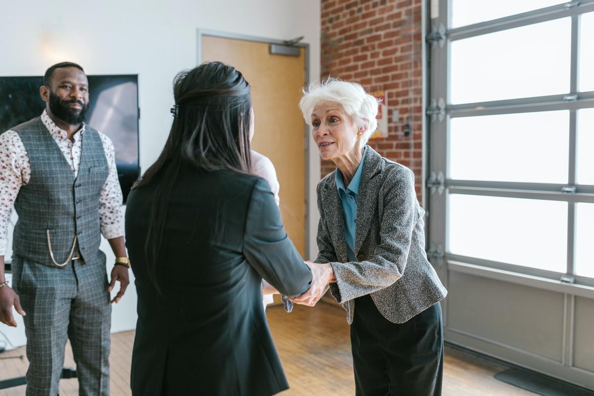An older woman with white hair in a grey blazer shakes hands with a woman in a black suit in a modern, industrial-style office