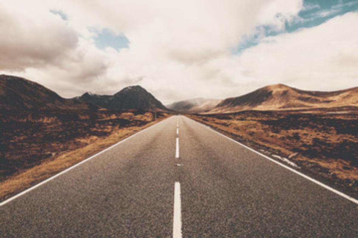 A long, straight asphalt road with a central white dividing line leading into the distance through a desolate, brown and yellow moorland or mountainous valley.