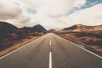 A long, straight asphalt road with a central white dividing line leading into the distance through a desolate, brown and yellow moorland or mountainous valley.