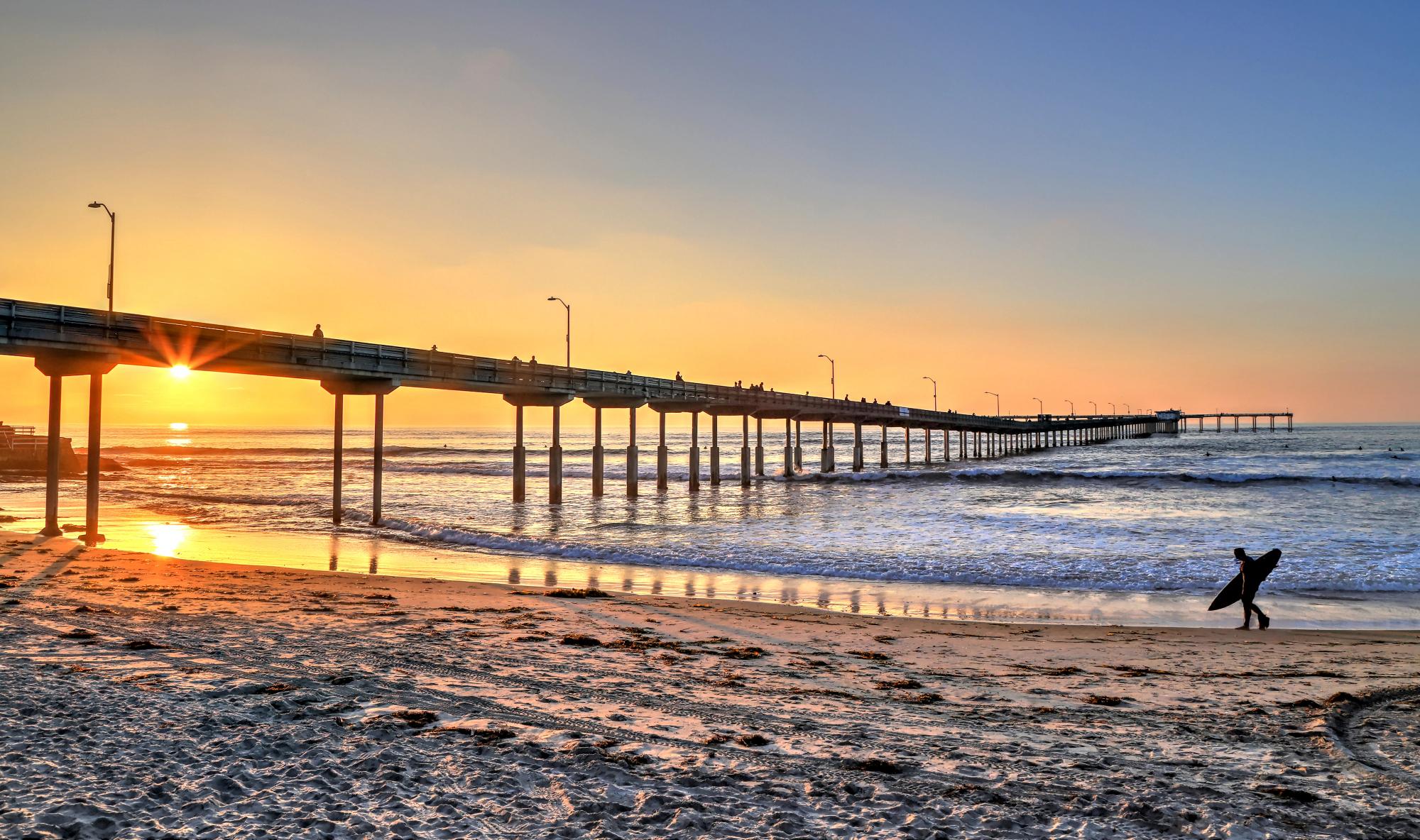 Ocean Beach Pier extending far into the Pacific Ocean during a golden sunset, with a lone surfer carrying a board walking along the sandy shore