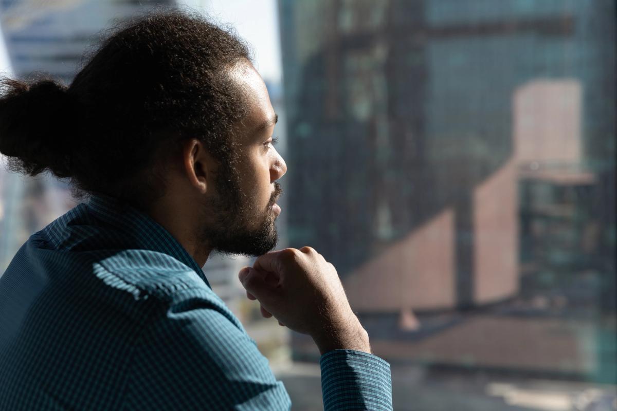 A thoughtful man with dark, curly hair tied back in a bun and a beard looks out a tall office window, resting his chin on his closed fist in a pensive pose.