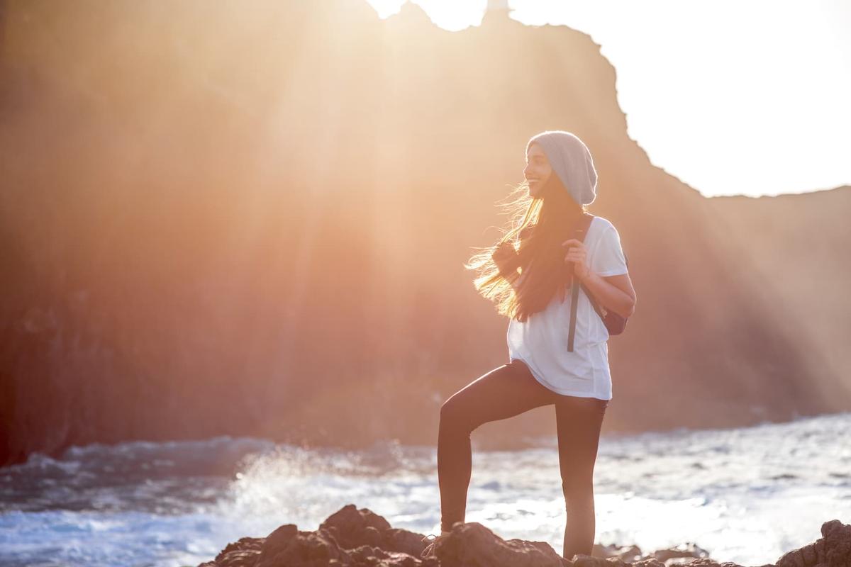 A woman stands on a rocky shoreline at the ocean’s edge, smiling into the distance with one foot raised on a rock. She wears a beanie, scarf, white t-shirt, leggings, and a backpack, while warm sunlight flares across the scene and waves crash behind her near a large cliff.