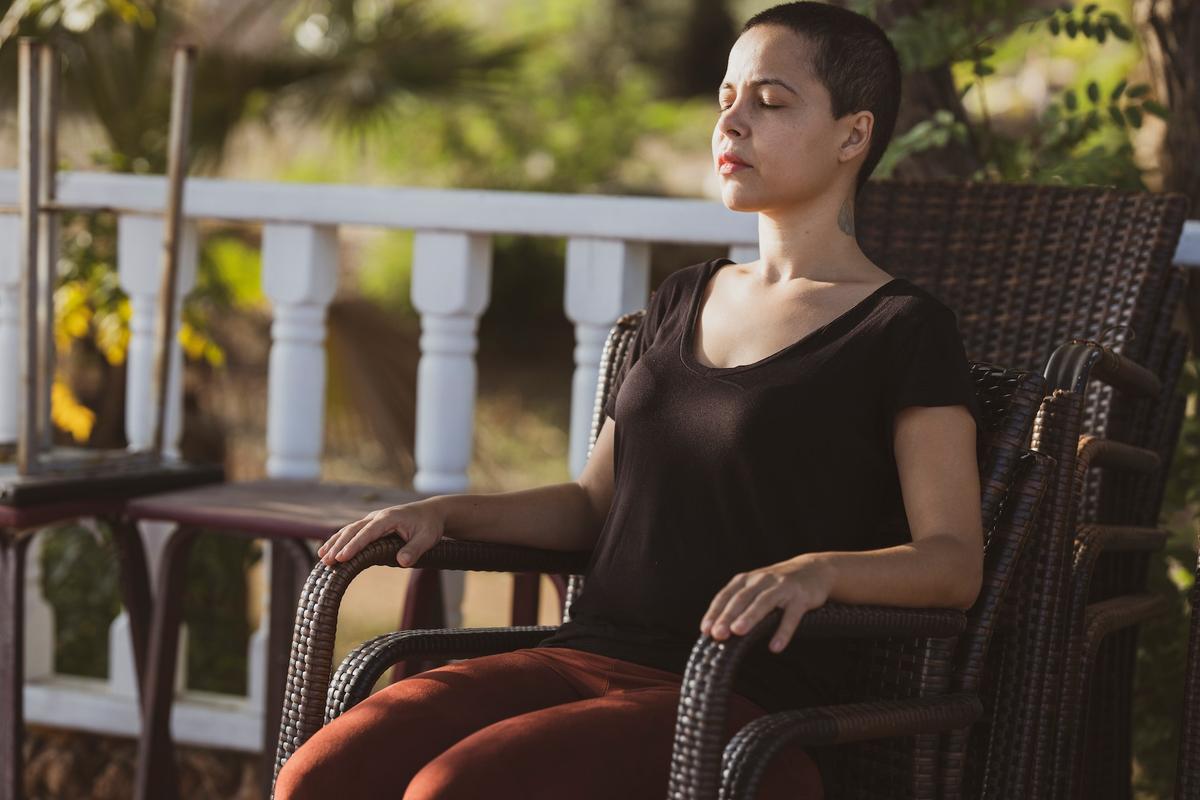 A woman with a buzzed haircut sits in a brown wicker chair on a porch, her eyes closed in a peaceful meditation.