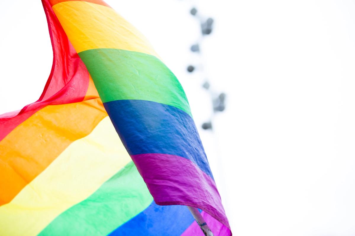 A close-up of a vibrant rainbow Pride flag billowing in the wind against a bright, overcast sky.