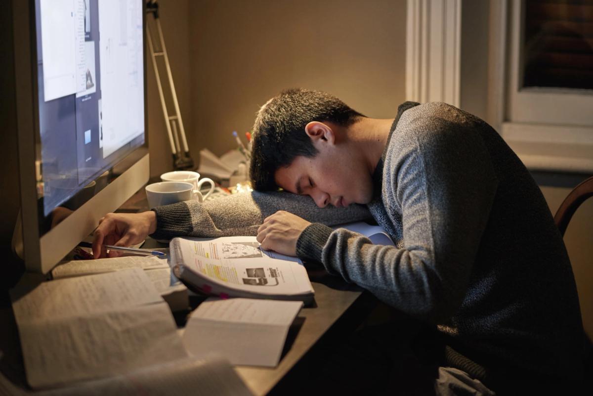 A young person, likely a student, is slumped over a desk asleep, resting their head on an open textbook surrounded by papers and a glowing computer screen, suggesting exhaustion from late-night studying or working.