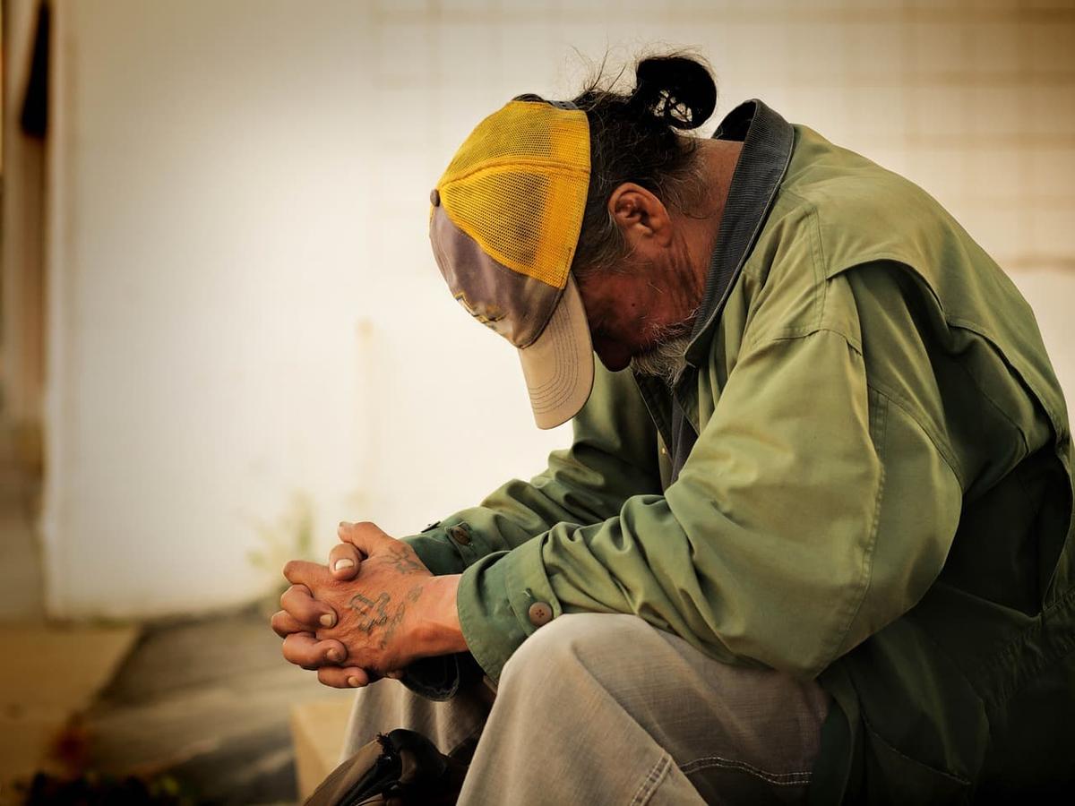A man sitting with his head bowed and hands clasped. He is wearing a green jacket and a yellow and grey trucker hat.