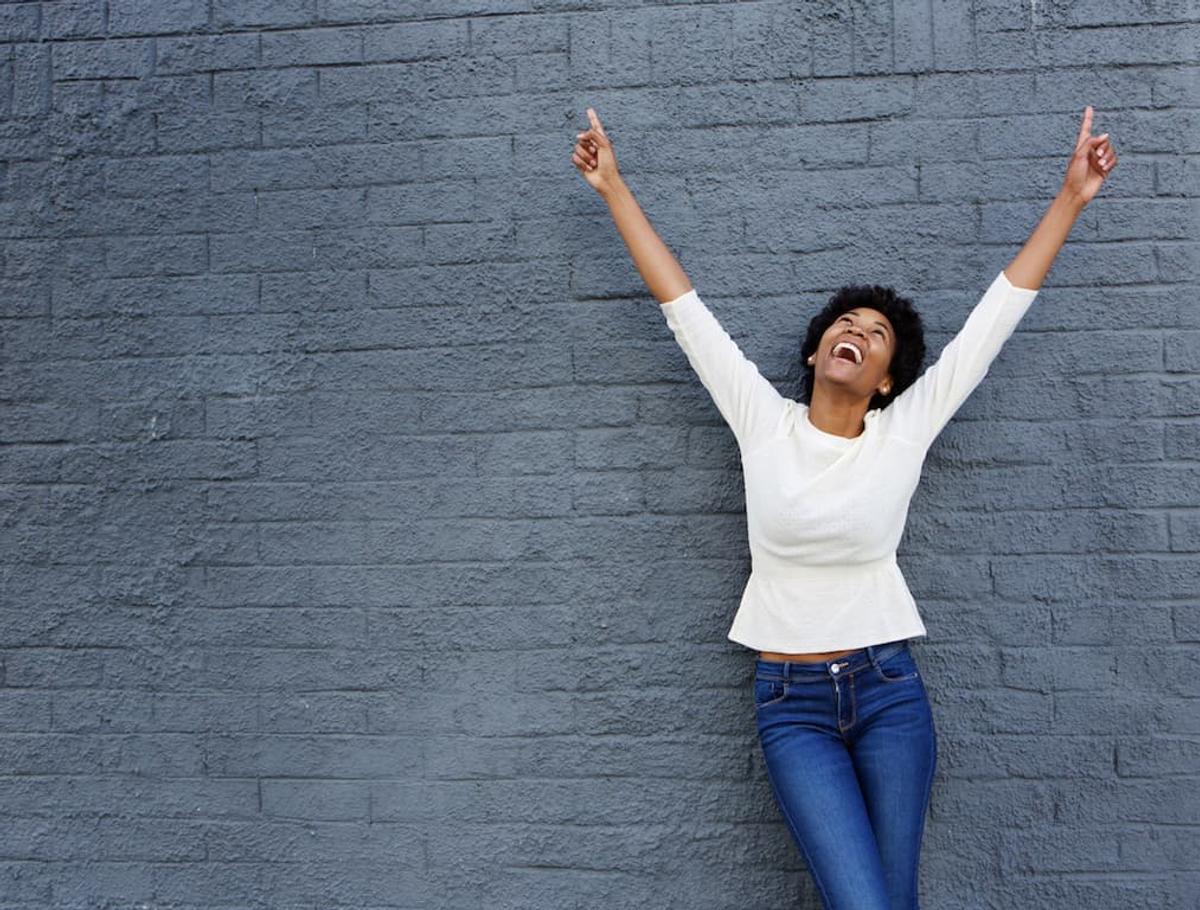 Smiling Black woman with arms raised in celebration against a gray brick wall.