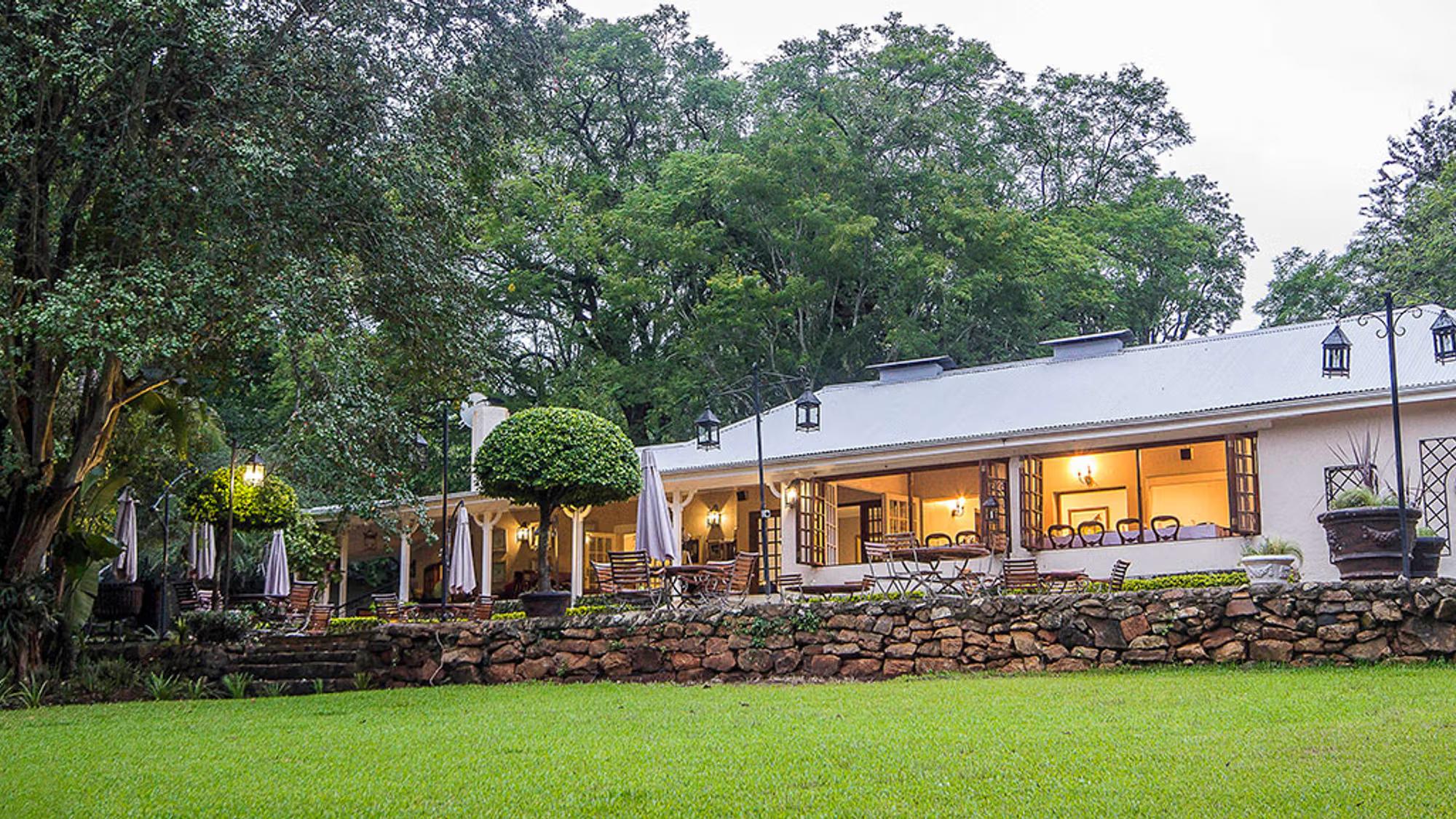 A long, single-story white house with a metal roof sits behind a rustic stone wall and a large green lawn.