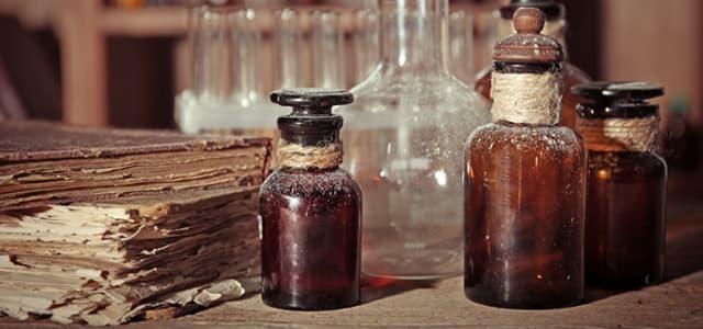 Vintage glass bottles filled with dark liquids sit on a wooden surface beside stacked old papers and laboratory glassware, evoking an antique apothecary or alchemy setting.