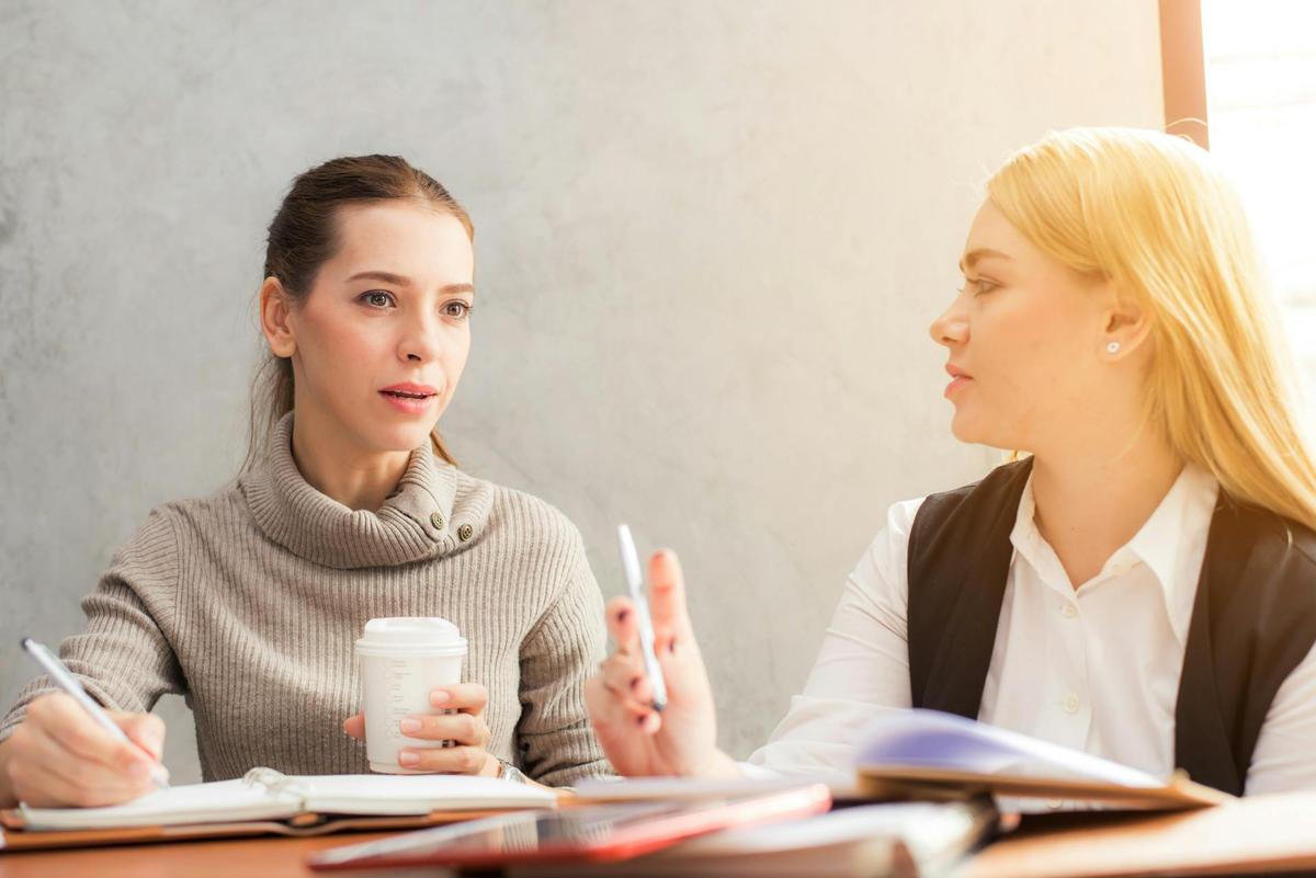 Two professional women in a brightly lit office setting, engaged in a focused discussion while taking notes and holding coffee.