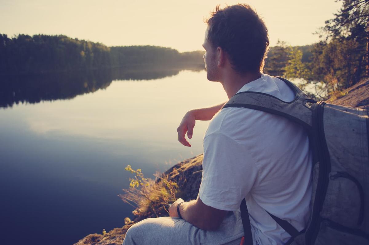 A man with a backpack sits on a rocky cliff overlooking a calm lake, silhouetted against the bright sunset or sunrise.