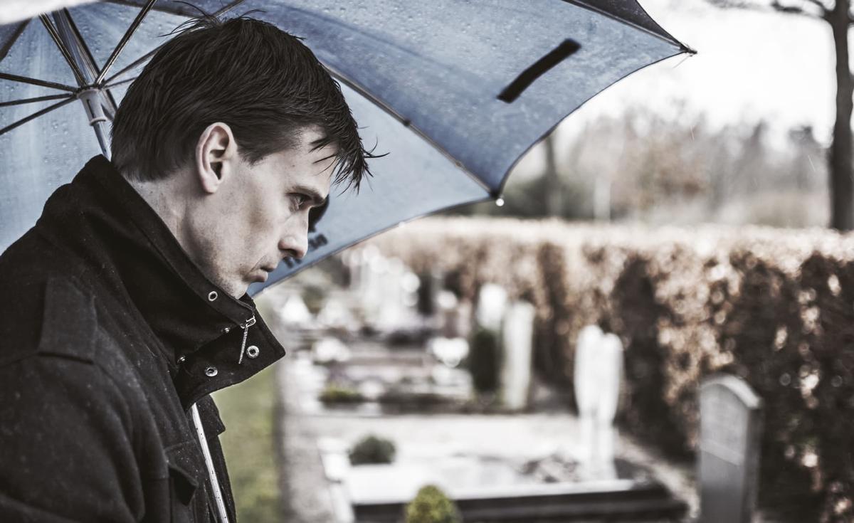 A somber man in a dark coat stands under a blue umbrella during a light rain, looking down with a mournful expression in a cemetery.