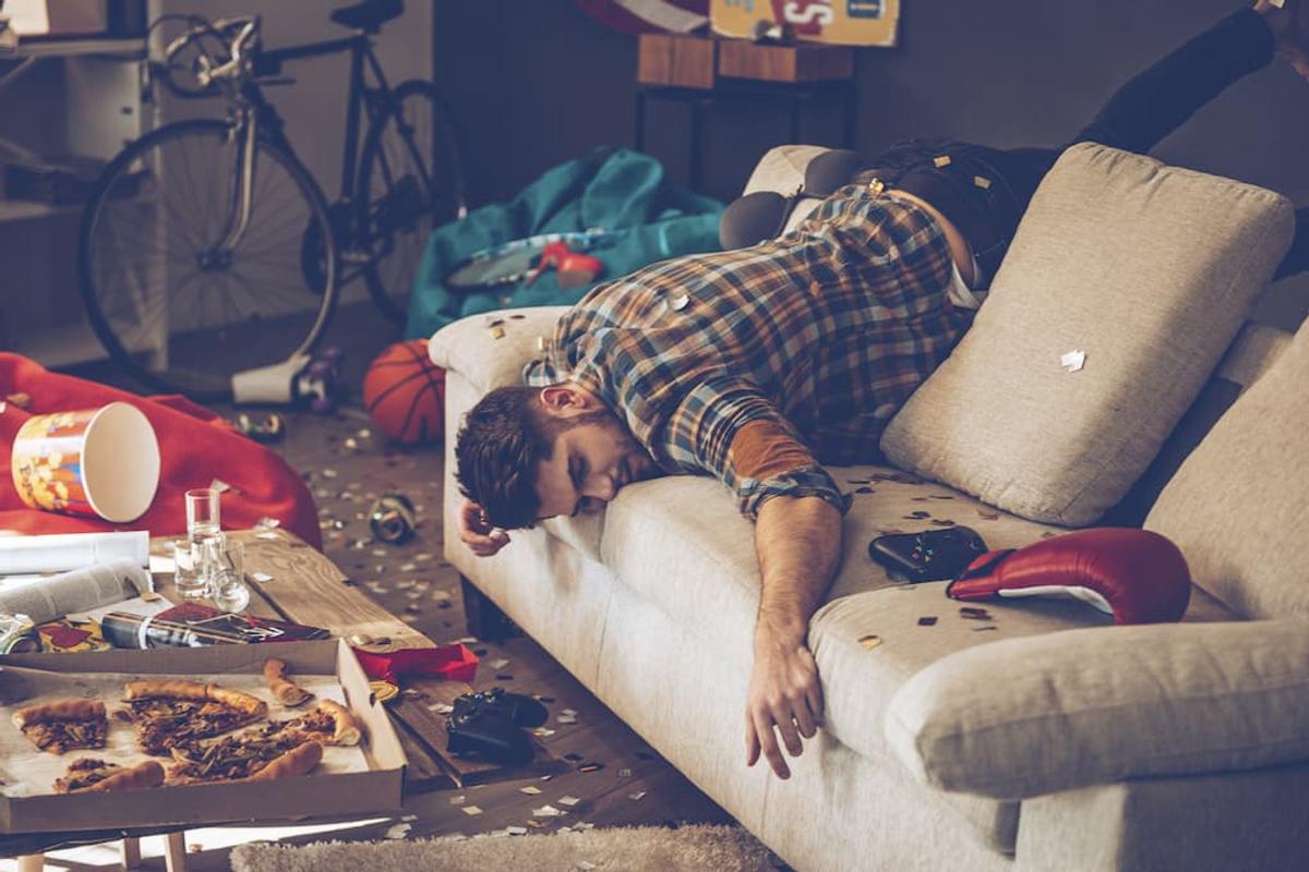A man lies face-down on a sofa in a very messy room filled with party remains and sports gear