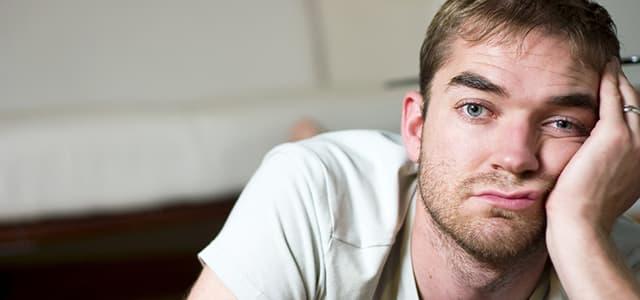 A close-up of a man with light brown hair and a slight beard, resting his head on his hand with a bored or weary expression.