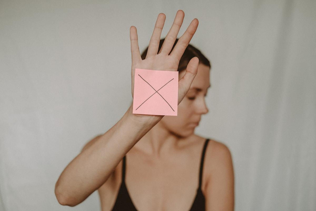 Woman holds up a hand in a “stop” gesture with a pink sticky note marked with an X, her face blurred in the background, symbolizing setting boundaries in recovery.