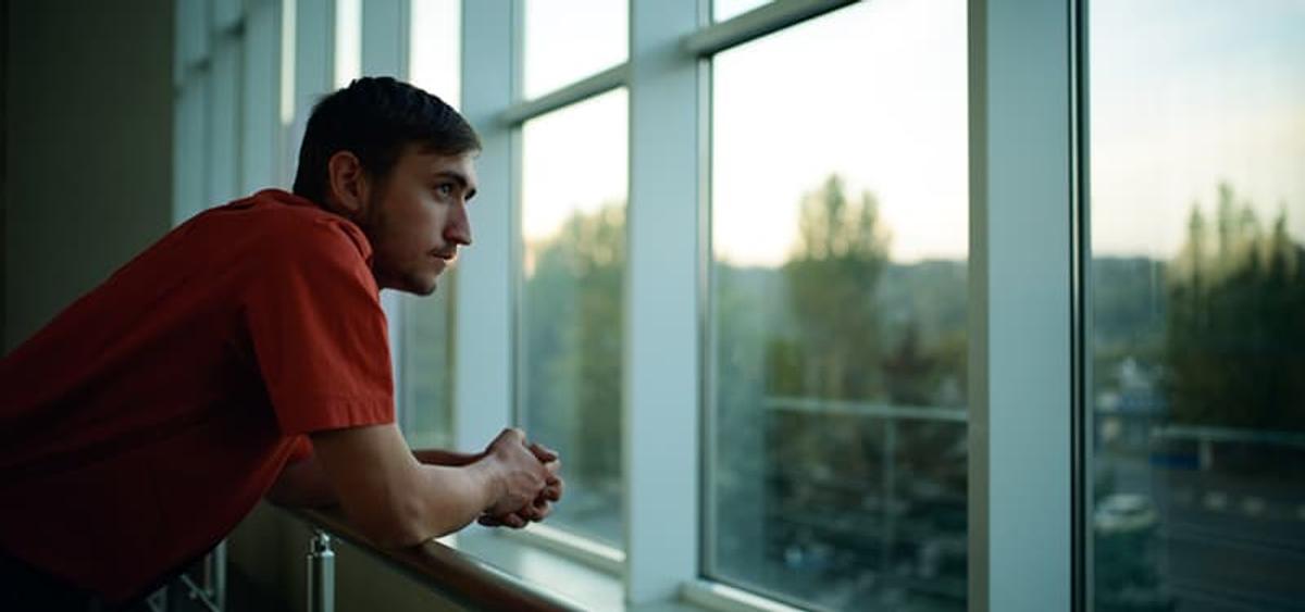A young man with dark hair and a red short-sleeve t-shirt is leaning on a metal railing, looking intently out a large, multi-paned window.