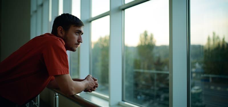 A young man with dark hair and a red short-sleeve t-shirt is leaning on a metal railing, looking intently out a large, multi-paned window.