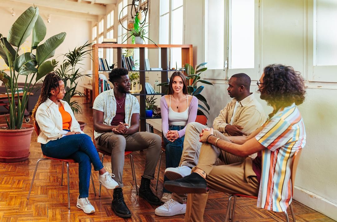 Group therapy session with diverse adults sitting in a circle, sharing experiences and offering support in a bright, welcoming counseling space