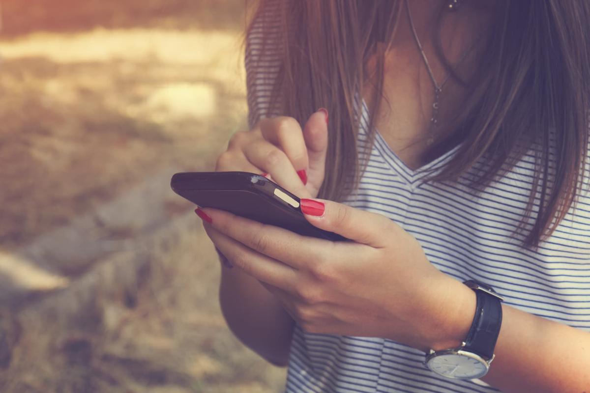 A close-up shot focusing on the hands of a person with long brown hair using a black smartphone outdoors.