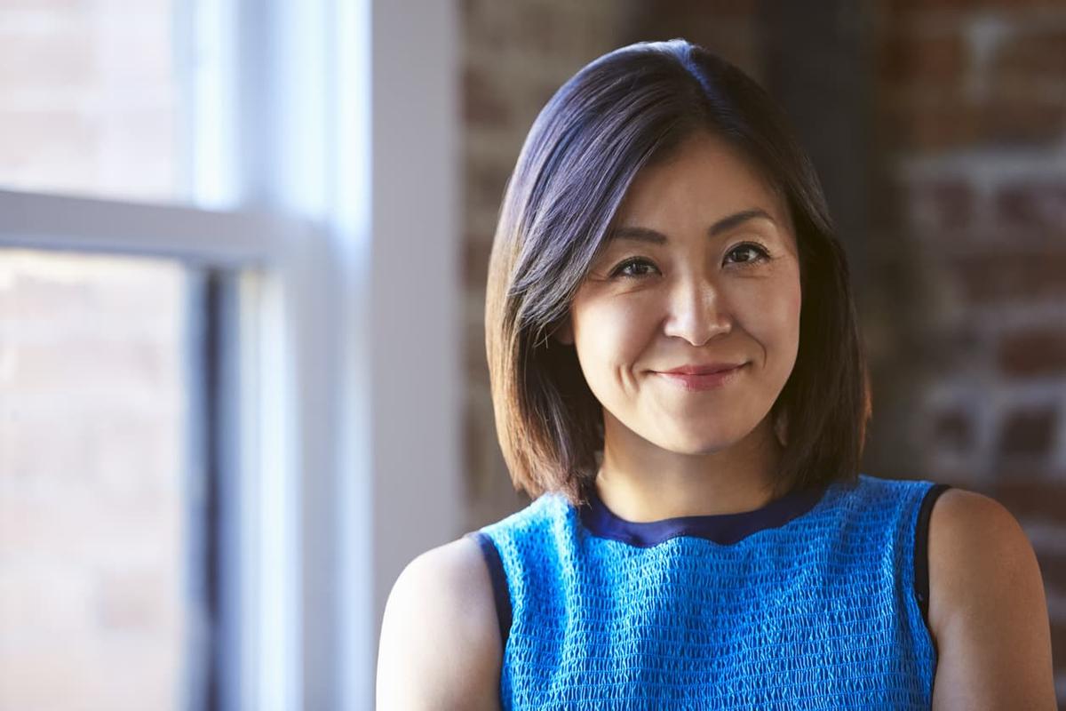 A close-up portrait of a smiling woman with shoulder-length dark hair wearing a textured blue top.
