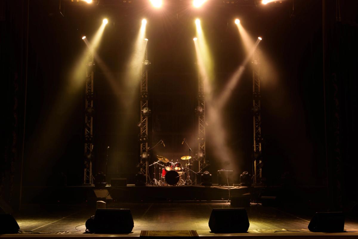 Stage set for a live music performance with a drum kit, microphones, and overhead stage lights.