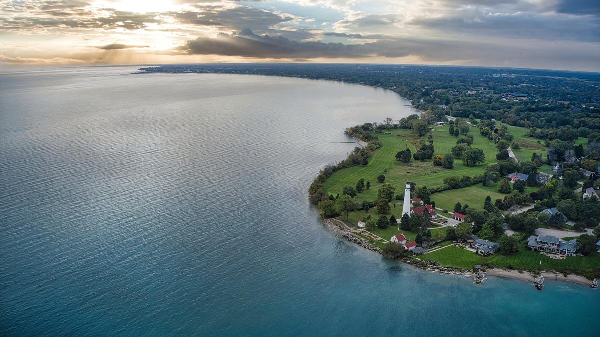 An aerial wide shot of a tall white lighthouse on a green coastal point, overlooking a vast body of water under a cloudy sunset.