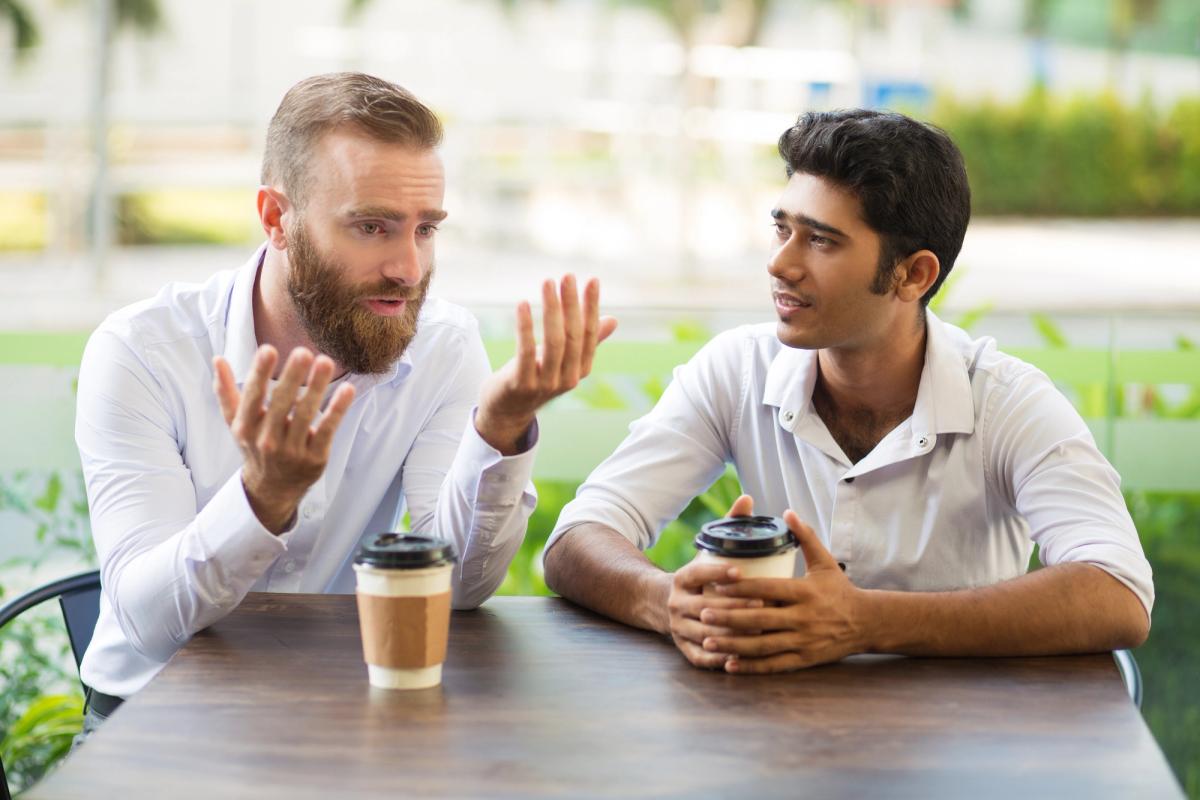Two men sitting at a table outdoors, holding coffee cups and having a thoughtful conversation, with one man gesturing as he speaks while the other listens attentively.