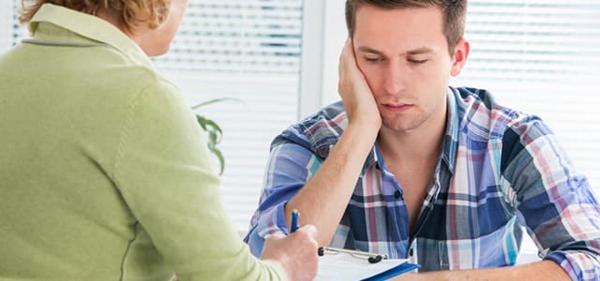Man sitting with his head resting on his hand while talking to a counselor who is holding a clipboard.