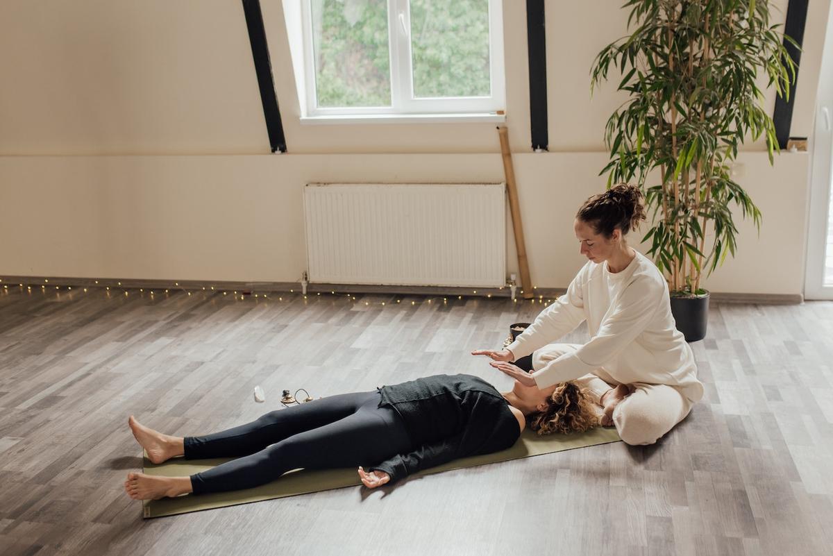 A person in a white sweatshirt sits cross-legged on a wooden floor, hovering their hands over another person lying in a relaxed pose on a yoga mat.