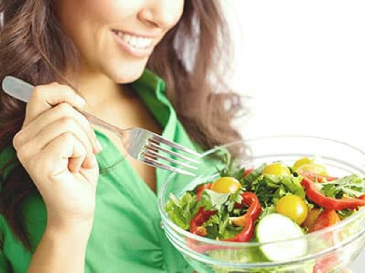 A smiling woman in a green shirt holds a silver fork over a clear glass bowl filled with a fresh garden salad containing lettuce, cucumber slices, cherry tomatoes, and red bell peppers.