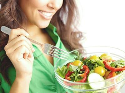 A smiling woman in a green shirt holds a silver fork over a clear glass bowl filled with a fresh garden salad containing lettuce, cucumber slices, cherry tomatoes, and red bell peppers.