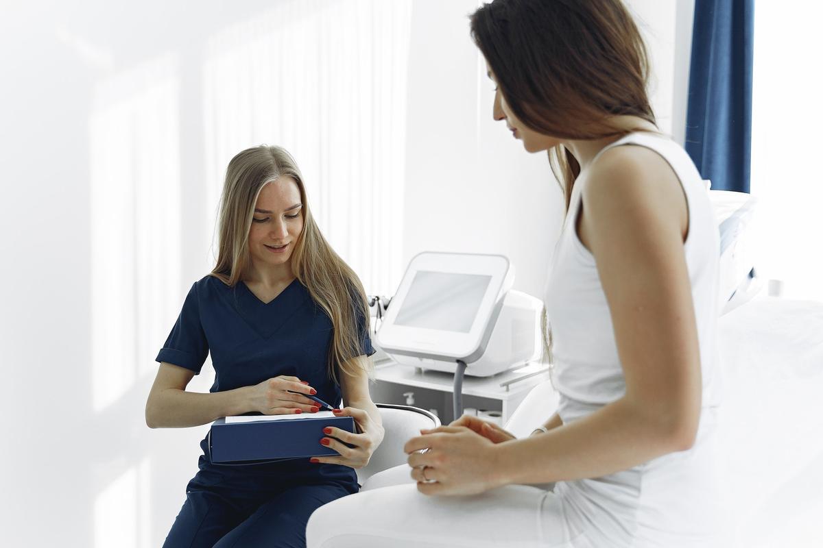 A healthcare professional in blue scrubs sits with a patient in a bright medical office, pointing to information on a clipboard during a consultation.