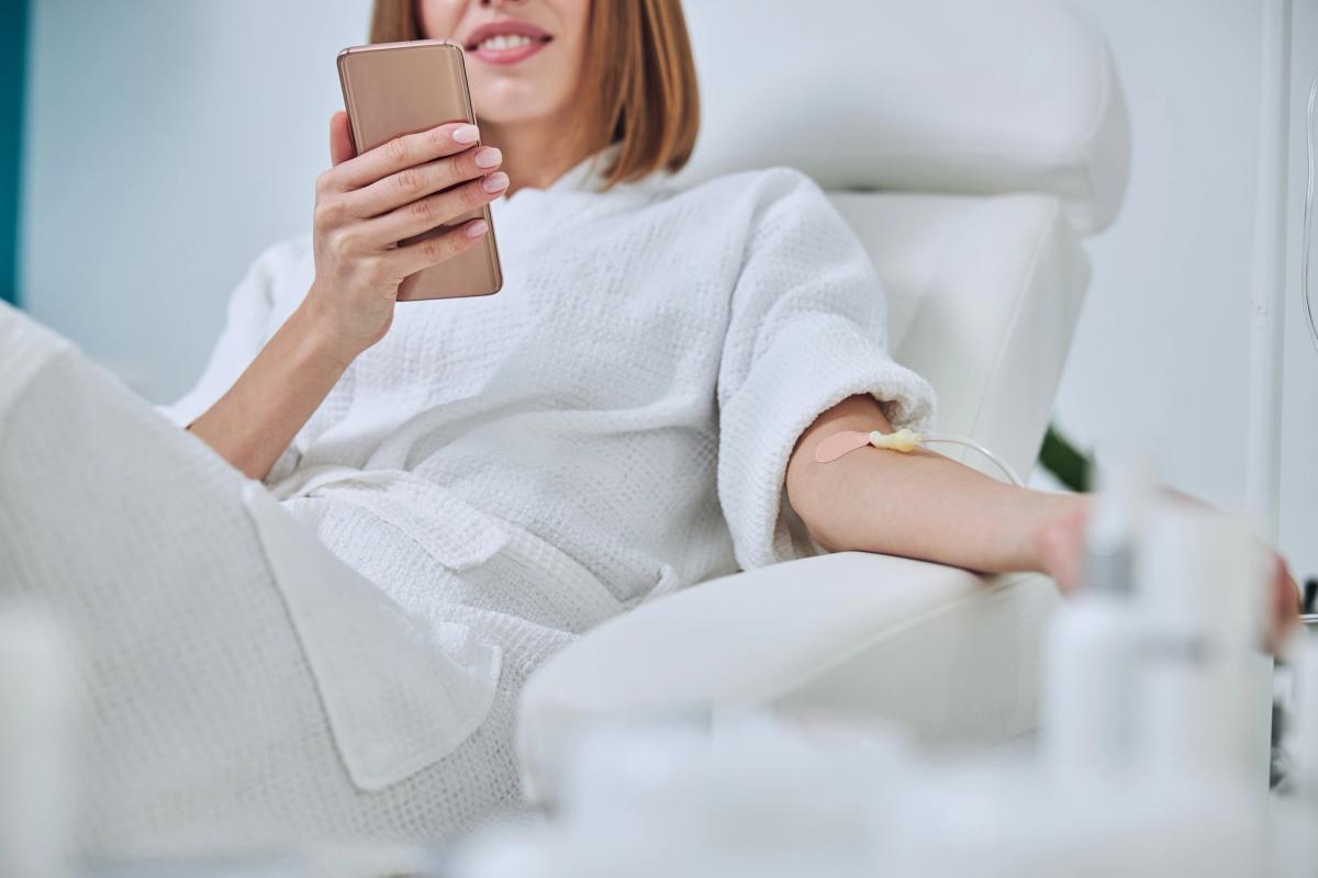 A woman in a white bathrobe sits in a clinical chair while receiving an IV drip and using her smartphone.