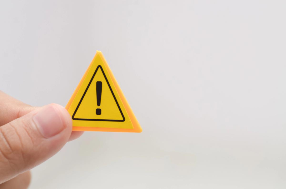 A hand holding a small, yellow triangular warning sign with a black exclamation mark against a plain light gray background.