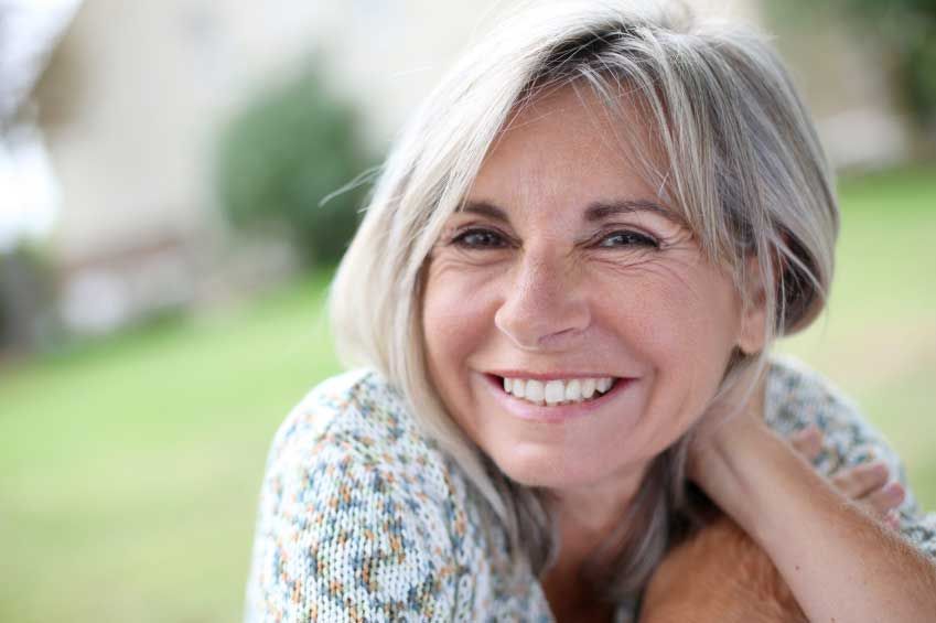 Close-up of a smiling woman with gray-blonde hair resting her head on her hand outdoors, with a blurred green background.