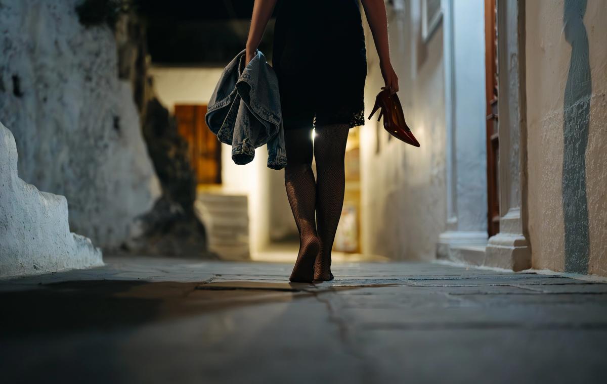 A woman walking barefoot down a narrow stone alley at night, carrying red high heels and a denim jacket.