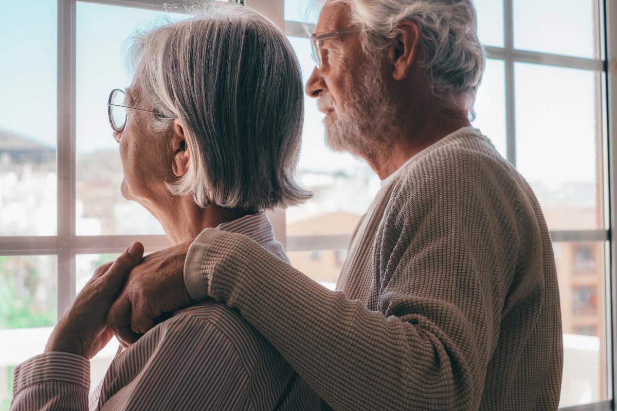 Elderly couple standing together by a window, the man gently resting his arm on the woman’s shoulder in support, representing how to help a loved one with addiction through compassion, patience, and steady encouragement.